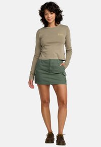 Lichtbruine cropped langemouwtop met gouden logo, gecombineerd met een groene cargorok met zijzakken en een label op de tailleband.