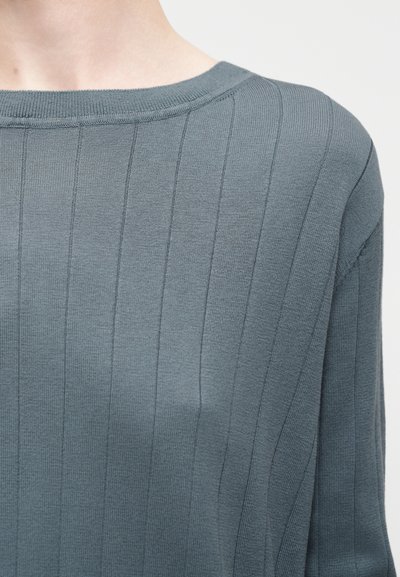 Κοντινό πλάνο ενός ατόμου που φοράει μπλε ριπ πλεκτό πουλόβερ με στρογγυλή λαιμόκοψη, εμφανίζοντας το ανώτερο μέρος του στήθους και των ώμων.