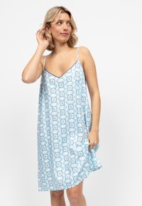 Donna con capelli biondi corti vestita con un abito senza maniche azzurro chiaro con un motivo geometrico a cerchi, che tiene il bordo dell'abito e guarda di lato.