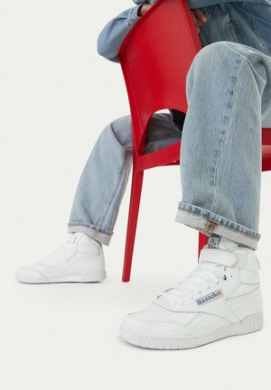 Person trägt hellblaue Jeans und weiße Reebok High-Top-Sneaker, sitzt auf einem roten Plastikstuhl vor weißem Hintergrund.