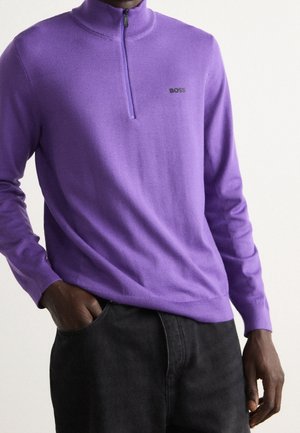 Uomo che indossa un maglione a zip corta viola con il logo BOSS e pantaloni neri, con una mano in tasca, su uno sfondo chiaro e semplice.