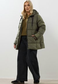 Doudoune vert olive avec capuche, dotée d'une fermeture éclair sur le devant, de cordons de serrage noirs et de poches latérales, assortie à un pull en tricot et un pantalon large noir.