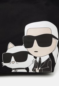 Czarna torba z nadrukiem przedstawiającym mężczyznę i białego kota, oboje w okularach przeciwsłonecznych. Płaska, teksturowana powierzchnia z konturowymi detalami.