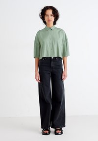 Camisa cropped verde claro com gola abotoada, combinada com calças largas pretas de cintura alta e sandálias pretas de tiras.