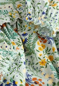 Tessuto a fantasia floreale in verde menta chiaro con bordi arricciati. Presenta fiori di vari colori tra cui giallo, blu e arancione su uno sfondo di fogliame verde.
