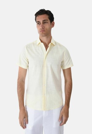 Uomo che indossa una camicia giallo chiaro a maniche corte con bottoni e pantaloni bianchi, in piedi davanti a uno sfondo chiaro e uniforme.