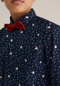 Modra majica z malimi belimi snežinkami in motivi Santa Klausa. Ima rdečo metuljčkasto kravato in ovratnik s gumbi. Gladka tekstura tkanine.