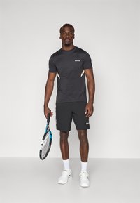 Svart idrottströja med ribbad textur och vit logotyp, tillsammans med svarta shorts med sidosträck. Håller en tennisklubba.