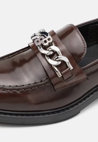 Mocassin en cuir marron avec une finition brillante, doté d'un accent chaîne argentée et d'un détail logo sur la bride. Semelle en caoutchouc noire.