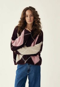 Grubo pletena pamučna puloverica u burgundiji, ružičastoj i krem boji s dijamantnim uzorcima. Dugi rukavi, široki manžeti, mekana tekstura. Uparen s plavim trapericama.