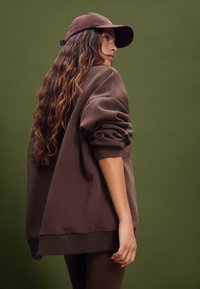Felpa oversize marrone con polsini a costine, abbinata a un berretto coordinato. Tessuto liscio, vestibilità rilassata e design minimalista.