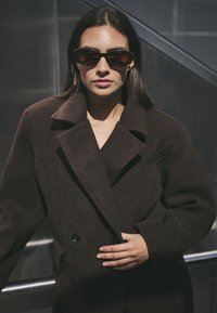Brūns divrindu uzvalks ar platu izgriezumu, izgatavots no teksturēta auduma. Akrila saules brilles ar elegantu, ovālu formu.