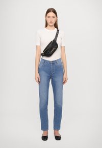 Črna prešita torbica na pas s zlatim logotipom, nosi se preko bele oprijete majice in hlač z visokim pasom v modri barvi, v kombinaciji s črnimi ravnimi čevlji.