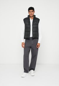 Melna pūkaina veste ar rāvējslēdzēju un augstu apkakli, uzvilkta virs baltas garām piedurknēm krekla, apvienota ar pelēkiem biksēm un baltiem sporta apaviem.