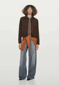 Bruine suède jas over een bruine gebreide trui, met een oranje gebreide ceintuuraccent; gecombineerd met een brede blauwe spijkerbroek.