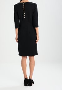 Vestido negro de largo hasta la rodilla con mangas de tres cuartos y corte recto. Presenta una fila vertical de botones en la parte trasera. Textura suave.