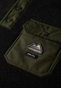 Sort fleece stof med en grøn nylon brystlomme. Lommen har et broderet bjerglogo og et "SUPERDRY®" skilt.