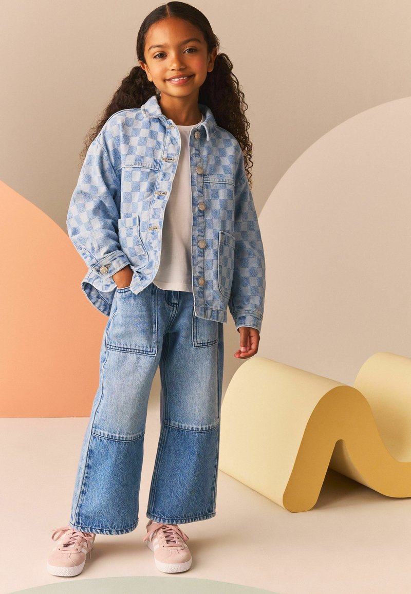 Veste en denim à carreaux bleu clair sur une chemise blanche, associée à un jean en denim à jambes larges. Des baskets roses complètent la tenue.