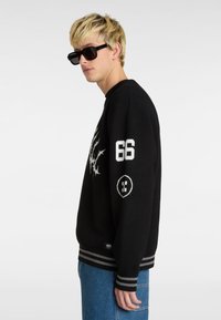 Suéter de punto negro con detalles gráficos blancos, incluyendo el número "66" y un logo, con puños y dobladillo de canalé a rayas grises.