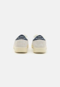 Fehér és szürke sneaker, puha velúrból készült sarokkal, kék akcentussal. A hátulján fehér "New Balance" logó található. Gumitalpak.