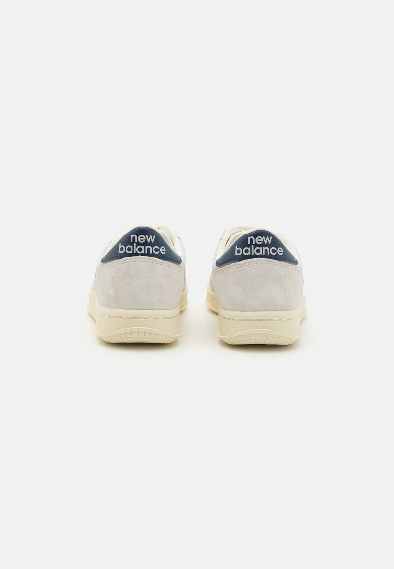 Zapatillas blancas y grises con un suave talón de ante y un detalle en azul. Logo "New Balance" en blanco en la parte posterior. Suelas de goma.