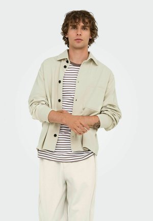 Chemise beige clair à boutons noirs sur un t-shirt rayé marron et blanc. Le modèle porte un pantalon couleur crème.