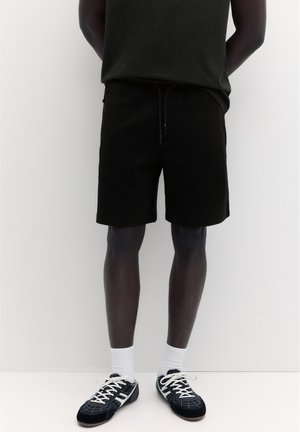 Personne se tient devant un fond blanc portant un short noir, un haut sombre, des chaussettes blanches et des baskets noires et blanches à lacets.