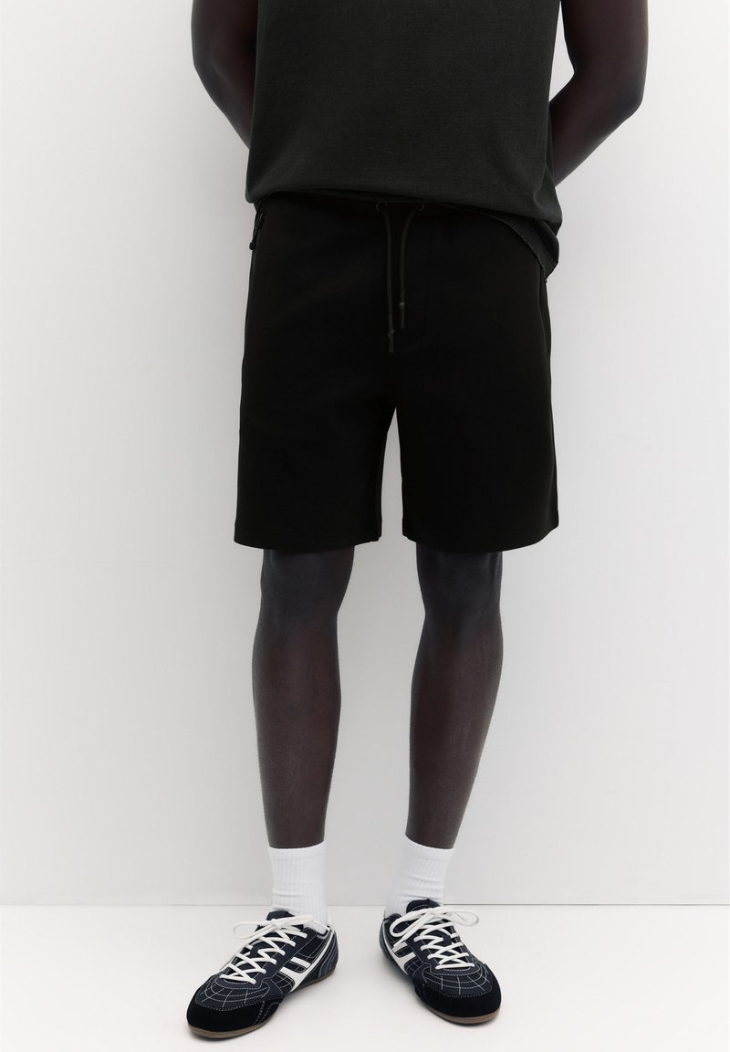 Personne se tient devant un fond blanc portant un short noir, un haut sombre, des chaussettes blanches et des baskets noires et blanches à lacets.