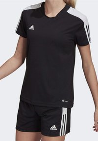 Kvinna iklädd svart Adidas sportställ med vita ränder på ärmarna och shortsen, står med armarna något utsträckta.