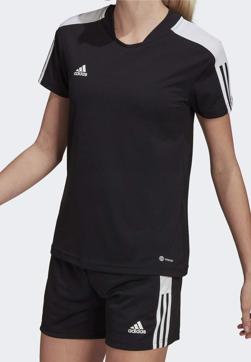 Kvinna iklädd svart Adidas sportställ med vita ränder på ärmarna och shortsen, står med armarna något utsträckta.