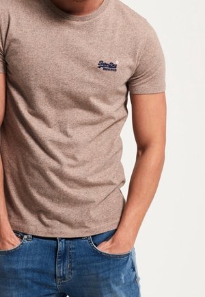 Man draagt een aansluitend beige T-shirt met korte mouwen, met een klein donker logo op de borst, en blauwe jeans, met de handen in de zakken.