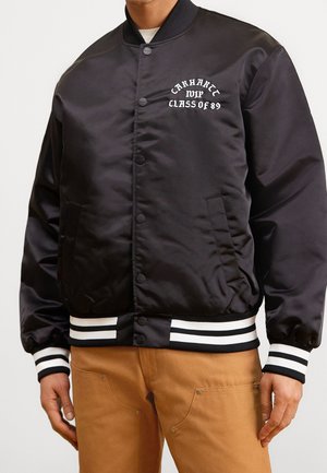 Czarna satynowa kurtka bomber z mankietami i dołem w białe paski, z haftowanym napisem "Carhartt WIP Class of 89" na lewej piersi, noszona z beżowymi spodniami.