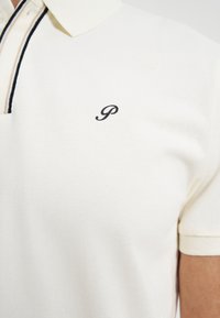 Polo blanc manches courtes avec col rayé noir et beige et lettre "P" noire brodée sur le côté gauche de la poitrine.