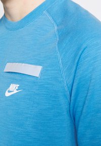Acercamiento de una persona que lleva una camiseta Nike de color azul brillante con el logo blanco y un pequeño bolsillo con solapa en el pecho.
