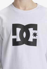 White long-sleeve shirt with a large black graphic featuring an intertwined "DC" logo and star accent. Soft cotton material, crew neckline.