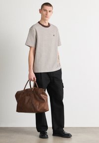 Pasiasta koszula z krótkim rękawem w kolorze beżowym i brązowym, połączona z czarnymi spodniami. Trzyma dużą brązową torbę skórzaną z dwoma uchwytami.