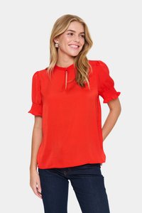 Blusa rossa con maniche a palloncino e dettagli di polsini smockati; presenta un colletto a chiave e un tessuto liscio; indossata su jeans scuri.