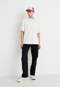 Weißes geripptes Polohemd mit kurzen Ärmeln, schwarze Hose und weiße Sneaker mit rotem Streifen. Dazu eine pink-weiße Trucker-Kappe.