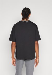 Camiseta de algodón negra con un corte relajado, hombros caídos y cuello redondo. La parte trasera del cuello presenta detalles decorativos. Jeans grises visibles.