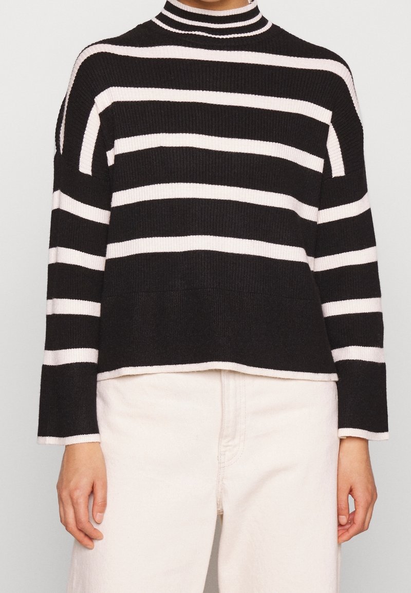 Osoba nosząca czarno-białą poziomo prążkowaną sweter dzianinowy z długimi rękawami i wysokim kołnierzem, w połączeniu z białymi spodniami.