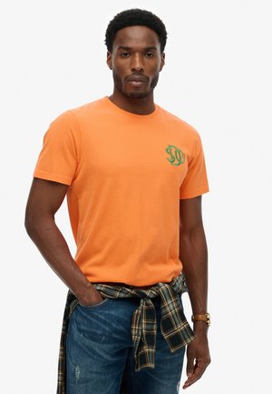 Mand iført orange t-shirt med grønt logo, blå slidte jeans og ternet skjorte bundet om livet, stående med den ene hånd i lommen.