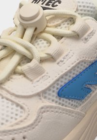 Krupan plan bijele tenisice s kremastim vezicama pričvršćenim okruglim kopčama za vezice i plavim logotipom ušivenim na mrežastom materijalu.