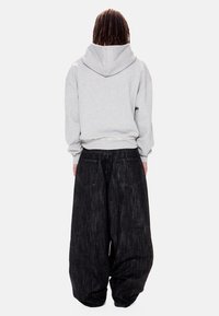 Άτομο με dreadlocks που φοράει ανοιχτό γκρι φούτερ με κουκούλα και υπερμεγέθη άνετο μαύρο τζιν, στέκεται με την πλάτη προς τα πίσω σε απλό λευκό φόντο.