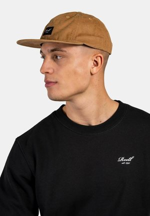 Braune Cordhut mit flachem Schirm, ausgestattet mit einem schwarzen Etikett auf der Vorderseite. Kombiniert mit einem schwarzen Sweatshirt, das einen runden Halsausschnitt hat.