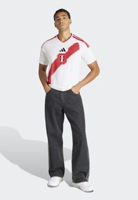 Witte jersey met korte mouwen, een rode diagonale streep en logo. Mesh textuur, V-hals ontwerp, gecombineerd met donkere wijde jeans en witte sneakers.