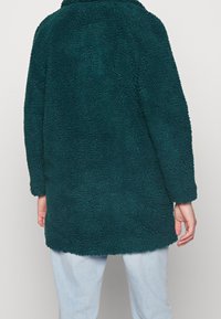 Teal, fluffy fleecejasje met een iets oversized ontwerp, lange mouwen en een ronde zoom. Geschikt voor casual draag.
