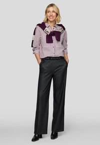 Lächelnde Frau steht mit den Händen in den Taschen, trägt ein gestreiftes Hemd mit Knöpfen, dunkle weit geschnittene Hosen, schwarze Stiefel und einen lilafarbenen Pullover lässig über die Schultern gelegt.