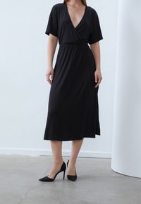 Mustaa kietaisumekkoa, jossa on lyhyet hihat, V-pääntie, vyötärön kiristys ja keskipitkä helma; yhdistetty mustiin, teräväkärkisiin korkokenkiin. Sileä kangaspinta.