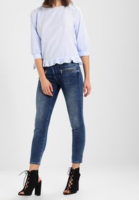 Haut rayé bleu clair avec un ourlet volanté, associé à un jean skinny bleu foncé et des bottes à lacets noires à talons hauts. Textures lisses tout au long.