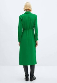 Grünes Langarmkleid mit Kragen, tailliertem Schnitt und knöpfbaren Manschetten. Glattes Material mit geradem Schnitt, kombiniert mit schwarzen Stiefeln.